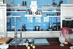 Kitchen island with sink and swivelling tap fittings; kitchen counter in background with white doors and reflective splashback
