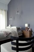 Chair with wooden back and ecru upholstered seat in front of double bed below pendant lamps with glass lampshades against grey wall