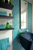 Black composite mineral sink and mirror on wall with turquoise mosaic tiles next to toiletries on floating shelves