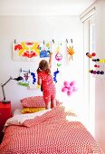 Little girl standing on bed in front of children's paintings on wall