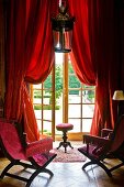 Antique chairs with pink velvet upholstery in front of French windows with red curtains and view into gardens