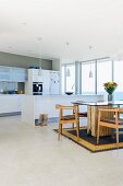 Offener Wohnraum mit moderner, weisser Küche mit Küchenblock; im Vordergrund ein gläserner Esstisch mit einem massiven Tischfuss und Holzstühlen auf einem Berberteppich