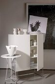 White crockery in designer cabinet with ornaments on top and vase on small side table in front of contemporary artwork