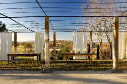 Rustic wooden tables beneath large pergola with wooden supports and white curtains against Mediterranean landscape