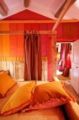 Gelb bezogene Kissen auf Himmelbett mit Holzgestell gegenüber Wand mit verschiedenen Rottönen in Streifenoptik in rustikalem Schlafzimmer