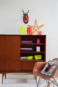 Hirschbüste mit Sicherheitsstreifen und zu Stern geformter Zollstock mit Kelchen und Kerzen in Neonfarben auf Retroschrank; Stuhlklassiker im Vordergrund