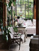 Gemütliche Verandaecke eines Landhauses - Rattanstuhl und rustikaler Holztisch mit abblätternder Farbe neben Tagesbett an Bleiglaswand