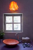 Atmospheric bathroom with ceramic wash basin and warm orange, hanging light over a pastel violet vanity in front of an exposed concrete wall