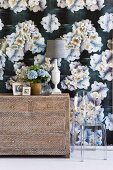 Bouquet of flowers and table lamp on a simple chest of drawers in front of a wall with dark wallpaper with a floral pattern