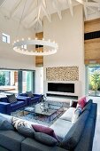 Large corner sofa and armchairs beneath a contemporary ring pendant light in a two story living room with hipped roof; ceiling height fireplace with firewood storage
