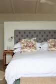 Double bed and vintage pillows in front of a gray upholstered headboard against a half-high room divider in a simple bedroom