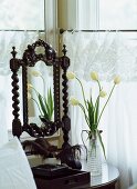 Weisser Tulpenstrauss in Glaskaraffe und antiker Schminkspiegel auf Tisch in Zimmerecke neben Fenster mit Spitzenvorhang