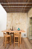 Bar table and matching wooden stools on wooden terrace below steel and wood balcony