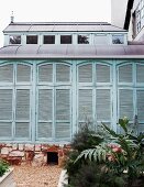 Extension with closed, turquoise shutters on stone foundation; raised flower beds and gravel paths