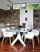 Dining area with Italian shell chairs at glass table; white cupboard against stone wall in background