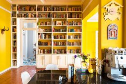 Gelber Wohnraum mit eingebauter Bücherwand; schwarzglänzender Tisch mit Stühlen und Kaffeemaschine im Vordergrund