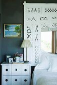White-painted bedside cabinet and decorative frame of perforated wood above bed