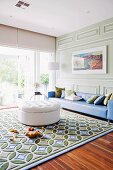 White ottoman on rug with graphic pattern and long blue couch against wall with classic, pastel green wood panelling