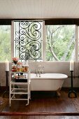Badewanne mit Klauenfüssen aus Messing vor Fensterfront mit Schmiedeeisengitter; Rollregal mit Badeutensilien