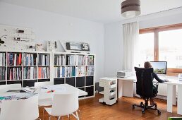 Weiß möbliertes Architekturbüro mit Frau an Computerarbeitstisch, Besprechungstisch und Modellen auf halbhoher Regalwand