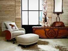 White armchair and matching footstool on animal-skin rug on floor next to rustic bench with leather seat cushion below window in wooden wall