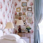 Corner of bedroom with table lamps and collection of pictures on wall covered in floral fabric