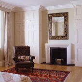 Elegant mirror above fireplace and English lounge chair in front of fitted wardrobe with panelled doors in bedroom