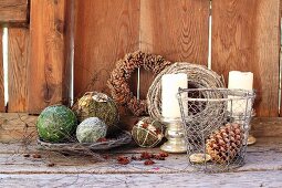 Advent arrangement of decorative spheres and wire basket of fir cones in front of candles and hand-made wreaths leaning on wooden wall