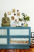 Buddha Figur auf blauem Sidebaord mit Glas Klapptüren vor Wand mit gerahmten Bildern