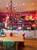 Küchenzeile an pinkfarbener Wand mit langen Holzregalen; davor ein einfacher Holztisch mit bunten Retro Stühlen