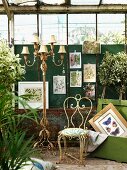Wrought iron chair and multiple-armed standard lamp in front of pictures of plants hung on wall in rusty greenhouse
