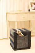 Square pouffe with hand-sewn, denim cover in front of rustic, white-painted console table with drawers