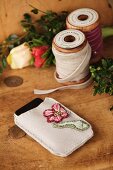 Embroidered mobile phone pouch and wooden reels of velvet ribbon on rustic wooden surface
