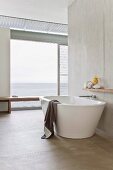 Free-standing, designer bathtub in purist bathroom in front of open glass wall with sea view