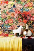 White horse ornament next to flowers in chrome vase against colourful, patterned wallpaper