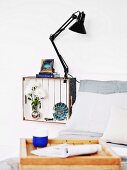 White wooden crate mounted on wall and painted white inside with ornaments and clip lamp used as bedside cabinet; breakfast tray made from crate in blurry foreground