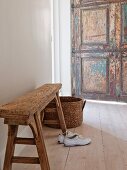 Trainers below rustic bench and rattan basket on wooden floor; weathered door in background