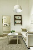 Weisser Couchtisch auf Teppich vor heller Sofakombination und schlichte Hängeleuchte in reduziertem Wohnzimmer, im Hintergrund Durchgang zur Küche