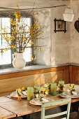 Dining area with place settings and floral arrangements below window; china jug of forsythia branches on windowsill