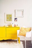 Gelbes Sideboard mit geschweiftem Sockel auf Flokatiteppich neben weißem Kunststoffstuhl, an der Wand weiße Bilderrahmen und ein Bild mit Frauenmotiv