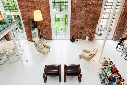 View from gallery into loft apartment with widely spaced armchairs and designer standard lamps against brick wall with floor-to-ceiling windows
