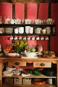 Dishes of vegetables and fruit on wooden dresser below crockery on wall-mounted shelves on red-painted wall