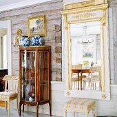 Antike Glasvitrine und vergoldeter Spiegel vor den rustikalen Wänden eines Blockhauses