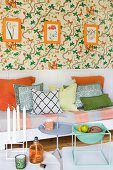 Bench with seat cushion and scatters cushions against white wall panelling below orange-framed botanical pictures on floral wallpaper