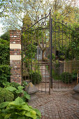 Open, wrought iron garden gate with brick gatepost and view of idyllic cottage surrounded by garden