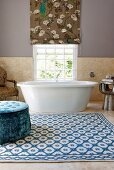 Spacious bathroom with blue velvet pouffe on patterned rug in front of bathtub below window