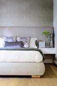 Gemütliches Doppelbett mit silberfarbenem Kopfende an grauer Wand; auf dem Bett silbergraue Zierkissen und eine Silbervase auf dem Nachttisch