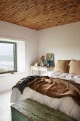 Simple, white bedroom with sea view, rustic bamboo ceiling and old wooden trunk at foot of double bed