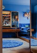 50s bar cabinet with mirrored doors and 70s, gilt chandelier above blue velvet sofa in living room with blue walls