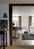 Delicate table lamp on console table next to wide, open doorway leading to living room with rococo footstool and armchairs